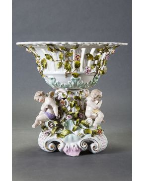 733-Gran centro de mesa en porcelana de Sajonia con marcas. Fuste con tres angelitos en bulto redondo que sostienen cesto calado con guirnaldas de flores 
